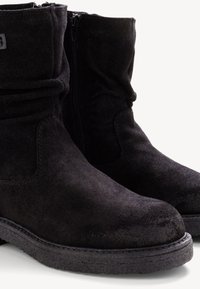 Stivaletti alla caviglia in suede nero con design arricciato, zip laterale, punta rotonda e suola texturizzata. Cuciture semplici accentuano i bordi.