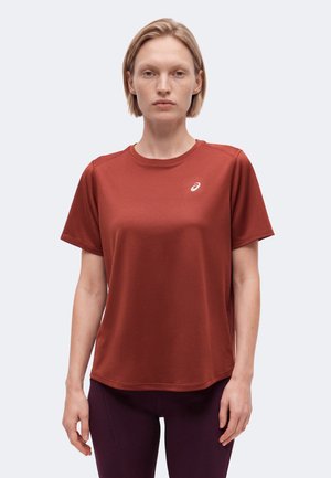 Femme debout, portant un t-shirt de sport à manches courtes de couleur rouille avec un logo blanc sur la poitrine et des leggings violet foncé.