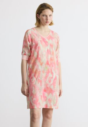 Γυναίκα που φοράει φόρεμα με κοντά μανίκια και λαιμόκοψη σε σχήμα V, με ροζ και μπεζ αφηρημένο σχέδιο, στέκεται μπροστά σε απλό φωτεινό φόντο.