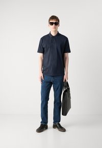 Camisa polo azul-marinho de manga curta, jeans, ténis pretos e óculos de sol escuros, segurando uma mala de couro preta contra um fundo neutro.