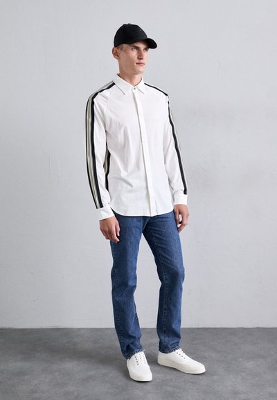 Vit långärmad skjorta med svarta randiga detaljer på ärmarna, i kombination med blå denimjeans och vita sneakers. Modellen bär en svart keps.