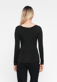 Kvinne med langt blondt hår som har på seg en svart langermet topp med blonde mansjetter og svarte mønstrede bukser, står vendt mot en hvit bakgrunn.