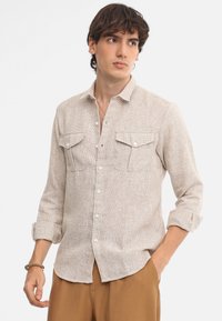 Camicia beige con bottoni realizzata in tessuto testurizzato, dotata di due tasche sul petto, bottoni frontali e maniche arrotolate.