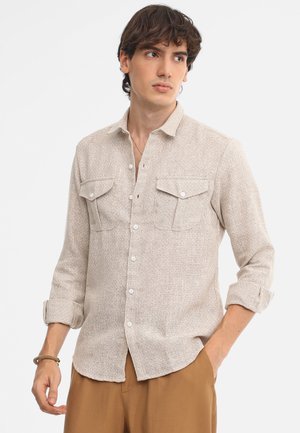 Camisa beige de botones hecha de una tela texturizada, con dos bolsillos en el pecho, botones en la parte delantera y mangas remangadas.