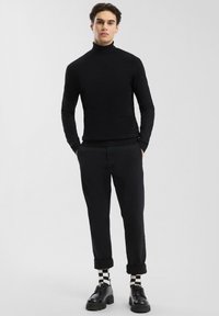 Jonge man in een zwart coltrui, zwarte opgerolde broek, zwarte schoenen en zwart-wit geruite sokken, staande tegen een effen lichte achtergrond.