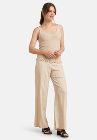 Beige gebreide tanktop met een ronde hals, gecombineerd met beige gestreepte wijde broek. Zwarte sandalen met bandjes maken de outfit compleet.