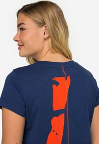 Camiseta azul marino hecha de tela suave, con un gráfico vertical en naranja brillante en la parte posterior, acentuado con costuras rojas a lo largo de la costura.