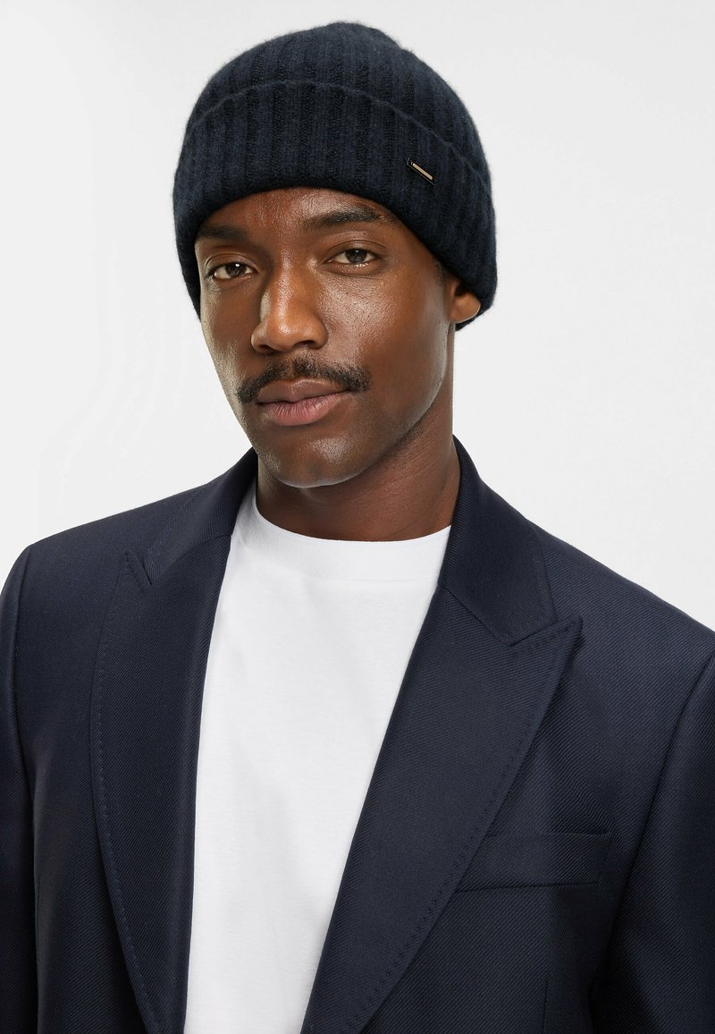Beanie a coste in navy scuro dal design elegante, con un sottile accento metallico. Indossato con una maglietta bianca e un blazer navy.