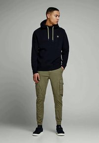 Jovem vestido com um hoodie preto com cordões amarelos, calças cargo verdes-oliva e sapatilhas pretas, posicionado contra um fundo cinza liso.