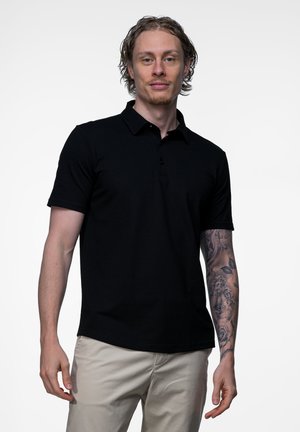 Jonge man met krullend haar draagt een zwart poloshirt en beige broek, toont een gedetailleerde tattoo-mouw op zijn rechterarm tegen een witte achtergrond.