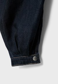 Mankiet ciemnoniebieskiej kurtki dżinsowej z beżowym przeszyciem i okrągłym metalowym zatrzaskiem na białym tle.