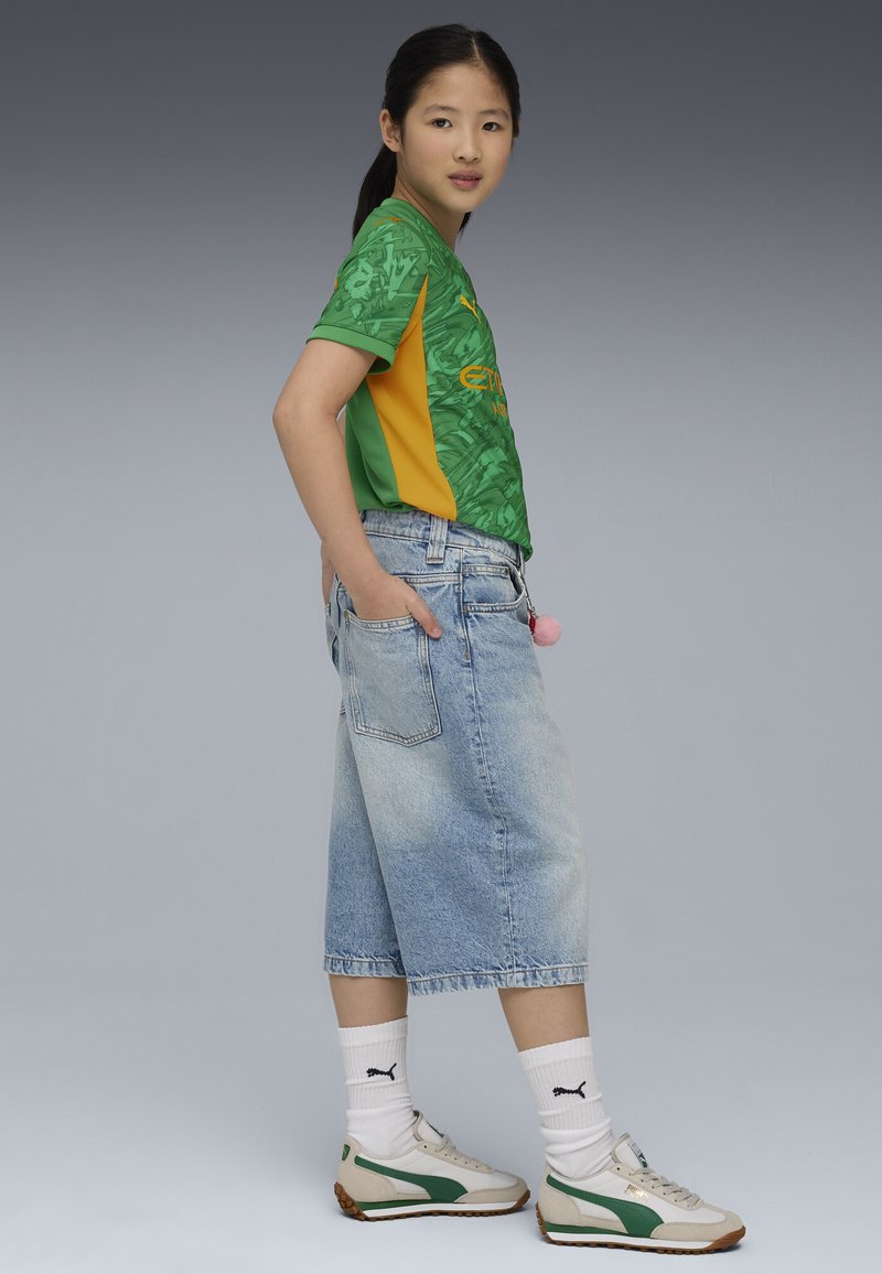 Camicia corta a maniche corte verde e gialla con design testurizzato, abbinata a culotte di jeans leggeri blu chiaro a vita alta e sneakers bianche con dettagli verdi.