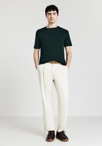 Uomo in piedi con le mani nelle tasche, indossa una camicia verde scuro a maniche corte, pantaloni bianchi con cintura marrone e scarpe marrone scuro su sfondo bianco.