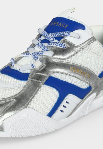 Baskets Versace blanches, bleues et argentées avec des motifs texturés, des lacets marqués et des logos Versace dorés sur la languette et le côté.