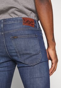 Vaqueros de mezclilla azul con una textura suave, que presentan una etiqueta de cuero marrón con "Lee" en la cinturilla y un único bolsillo trasero.