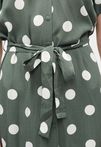 Grünes Kleid mit großen weißen Tupfen, vorne mit Knöpfen und einem Stoffgürtel, der in einer Schleife an der Taille gebunden ist.