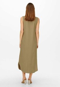 Donna con capelli castano chiaro fino alle spalle, indossa un abito midi verde oliva senza maniche e sandali con tacco trasparente, in piedi di spalle.