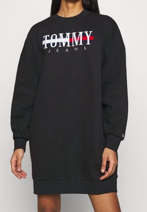 Kobieta w czarnej, oversize sukience typu sweatshirt Tommy Jeans z długimi rękawami i logo w kolorach białym, czerwonym i niebieskim na piersi.