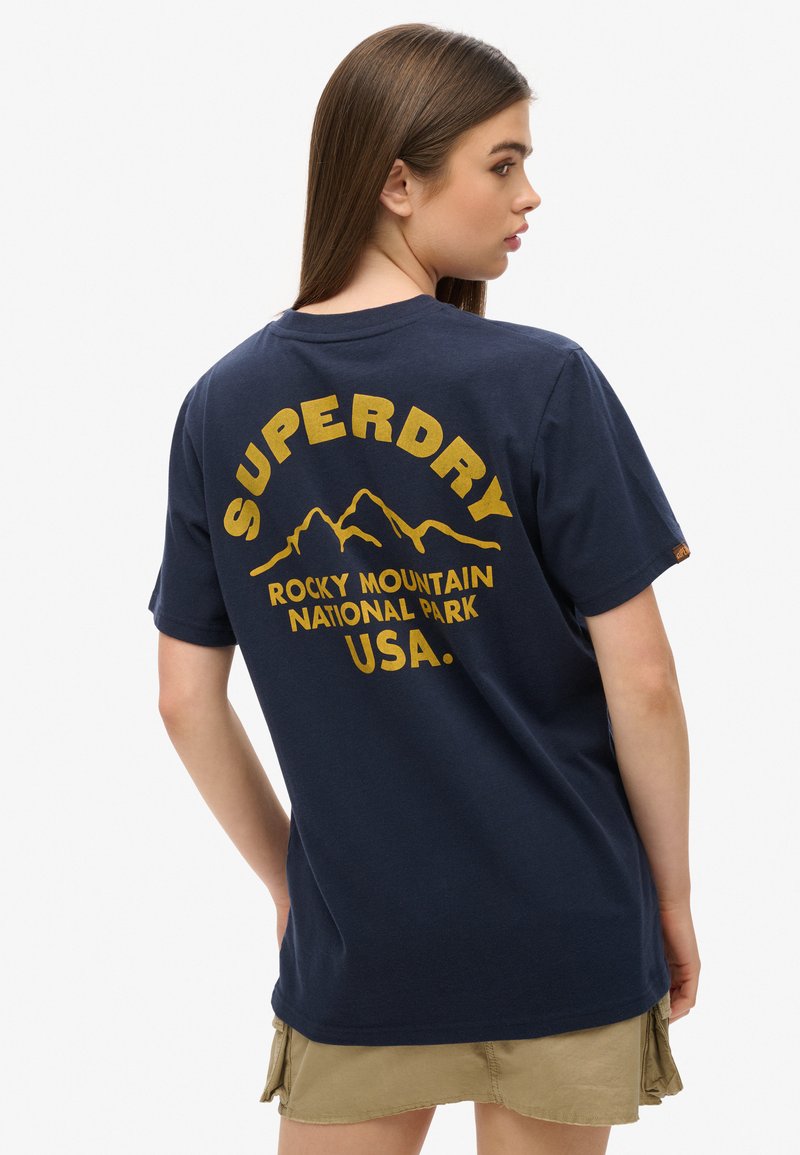Femme aux longs cheveux bruns portant un T-shirt bleu marine avec le texte jaune "Superdry Rocky Mountain National Park USA" et un graphisme de montagne au dos.