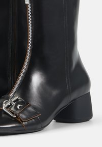 Botas de tobillo de cuero negro con acabado brillante, cremallera lateral, punta cuadrada y una hebilla decorativa en la parte frontal. Tacón robusto para mayor estabilidad.