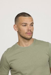 Un homme avec les cheveux courts et une légère barbe portant un t-shirt vert sauge, regardant légèrement vers la droite sur un fond clair et uni.