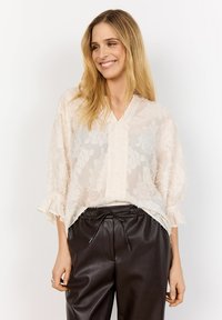 Leende kvinna med långt blont hår som bär en genomskinlig cremefärgad blus med blommönster och svarta läderbyxor med dragsko, mot vit bakgrund.