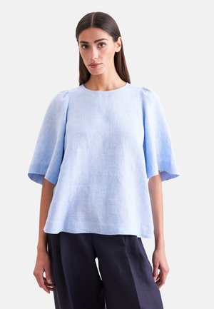 Kobieta z prostymi ciemnymi włosami nosi jasnoniebieską bluzkę z krótkim rękawem, szerokimi rękawami i luźnym krojem, zestawioną z ciemnymi spodniami.