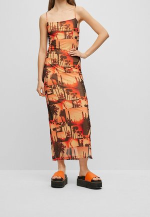 Femme portant une robe maxi ajustée avec des imprimés coucher de soleil désert orange et marron et cactus, associée à des sandales compensées noires avec des lanières orange.