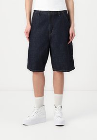 Mörka denimshorts med avslappnad passform, som har framfickor, minimala sömnadsdetaljer och en ofärdig nederkant, i kombination med vita sneakers.