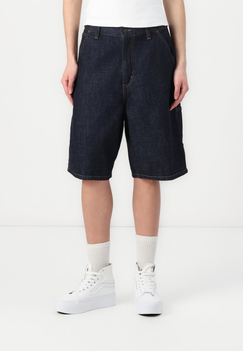 Mörka denimshorts med avslappnad passform, som har framfickor, minimala sömnadsdetaljer och en ofärdig nederkant, i kombination med vita sneakers.