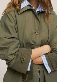 Personne portant un trench-coat vert boutonné par-dessus une chemise rayée bleue et blanche, les bras croisés sur la poitrine.