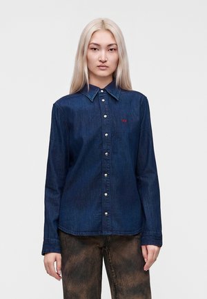 Jonge persoon met lang blond haar die een donkerblauw denim blouse met knopen en vervaagde bruine broek draagt, staand tegen een effen lichte achtergrond.