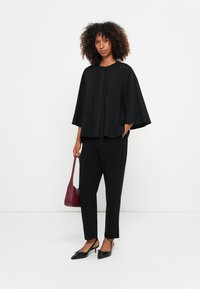 Mulher vestindo uma blusa preta larga com mangas amplas, calças pretas, sapatos de salto agudo pretos, e segurando uma bolsa bordô contra um fundo branco.