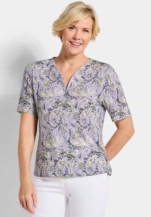 Blonde vrouw die een paars shirt met paisley-patroon en korte mouwen draagt, met een rits aan de voorkant, gecombineerd met een witte broek, glimlacht met de hand achter haar rug.