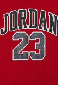 Rød t-shirt med "JORDAN" i store grå bogstaver over "23," med et tekstureret, broderet design mod en glat stofbaggrund.