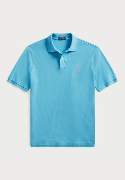Cămașă polo de un albastru deschis, fabricată din bumbac. Prezintă un gât cu trei butoane, mâneci scurte și un mic logo roz pe pieptul stâng. Textură netedă.