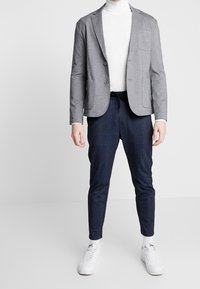 Blazer grigio con due tasche frontali, abbinato a pantaloni a quadri blu navy con cintura elastica. Un dolcevita bianco e sneaker bianche completano l'outfit.