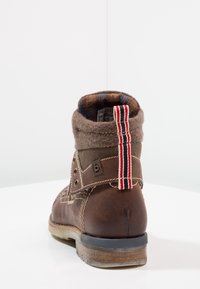 Bota de cuero marrón con un puño de lana gris, que presenta una lengüeta de tela a rayas y ojales de metal. Superficie texturizada y suela duradera.