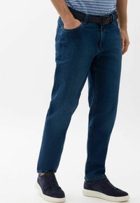 Jean en denim bleu foncé avec une coupe droite, cinq poches et une ceinture noire. Assortis à des baskets navy avec des semelles blanches.