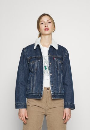 Giacca di denim con colletto in shearling, colore blu scuro, chiusura con bottoni, tasche laterali e dettagli di cucitura sottili, indossata sopra una t-shirt grafica.