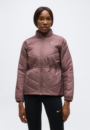 Kvinde iført en mauve farvet quiltet jakke med lynlås og sorte leggings med hvide Nike-logoer, stående foran en ensfarvet hvid baggrund.