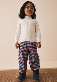 Pull blanc en maille torsadée, pantalon à carreaux violet et jaune, et bottines bleues avec des pampilles. Motifs texturés sur l'ensemble de la tenue.