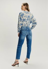 Kobieta stojąca z ręką na biodrze, ubrana w niebieską kwiecistą bluzkę, niebieskie dżinsy i czarne sandały na obcasie na jednolitym jasnym tle.