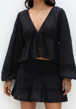 Kobieta ubrana w czarną, długorękawową, zapinaną na guziki bluzkę oraz dopasowaną, warstwową mini spódnicę z teksturowanymi detalami.