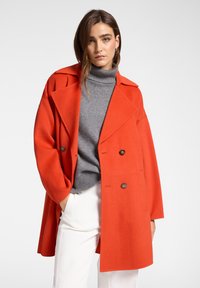 Cappotto di lana rosso dal design doppiopetto, con ampi risvolti e dettagli con bottoni, indossato sopra un dolcevita grigio e pantaloni bianchi.