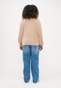 Μπεζ φλις πουλόβερ με μακριά μανίκια, σε συνδυασμό με ανοιχτό μπλε παντελόνι φαρδιάς γραμμής, εμφανίζεται από πίσω σε έναν απλό λευκό φόντο.