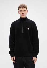 Čierny pletený sveter s vysokým golierom a polovičným zipsom. Na hrudi má malú červenú a bielu logovú nášivku. Textúrovaný, s voľným strihom.