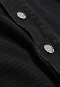 Tissu en denim noir avec deux boutons-pression métalliques argentés gravés d'un texte sur une partie pliée du matériau.