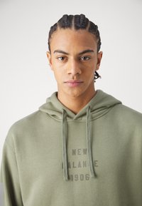 Sudadera verde oliva con cordones, que presenta un estampado frontal que dice "NEW BALANCE 1906" en un tono más oscuro. Tejido suave y texturizado.
