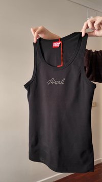 Mains tenant un débardeur noir Diesel avec un logo en strass sur le devant et une étiquette rouge attachée, fond de mur uni.